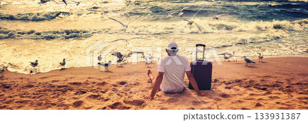 A human sits on the beach in summer with a travel suitcase. The man looks at the sea. Seagulls fly over the beach 133931387