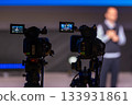 Two professional video cameras on tripods film a speaker on stage during a live presentation 133931861