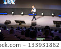 Confident presenter on a bright stage addressing a seated audience during a conference or seminar 133931862