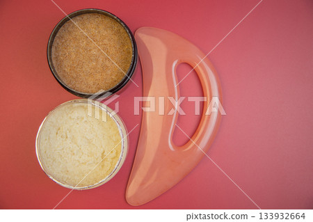 Two jars of body scrubs, one brown and one cream, alongside a pink massage tool on a red background, highlighting beauty and self-care essentials Two jars of body scrubs, one brown and one cream, alongside a pink massage tool on a red background, highlighting beauty and self-care essentials 133932664