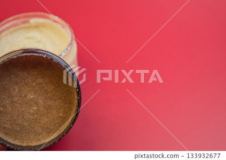 Natural body scrub jar reveals creamy brown texture, set against a vivid red backdrop, highlighting beauty, skincare, and self-care themes in a visually appealing manner Natural body scrub jar reveals creamy brown texture, set against a vivid red backdrop, highlighting beauty, skincare, and self-care themes in a visually appealing manner 133932677