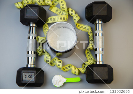Black dumbbells are positioned alongside a measuring tape and a protein powder container, showcasing fitness essentials for a workout routine and healthy living Black dumbbells are positioned alongside a measuring tape and a protein powder container, showcasing fitness essentials for a workout routine and healthy living 133932695