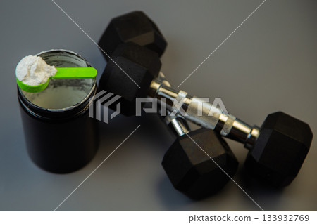 Set of black dumbbells alongside a protein powder container with a green scoop, placed on a gray surface, emphasizing fitness and nutrition essentials for workouts Set of black dumbbells alongside a protein powder container with a green scoop, placed on a gray surface, emphasizing fitness and nutrition essentials for workouts 133932769