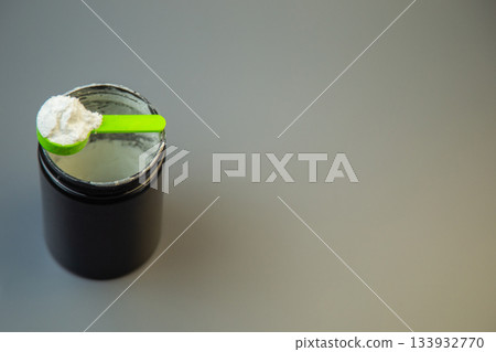 Black jar filled with white powder and green scoop on gray surface, representing nutritional supplement for fitness and health, perfect for dietary needs and wellness goals Black jar filled with white powder and green scoop on gray surface, representing nutritional supplement for fitness and health, perfect for dietary needs and wellness goals 133932770