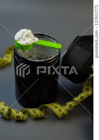 Black protein powder container features green scoop on top, surrounded by yellow measuring tape and dumbbells, representing fitness and health goals 133932772