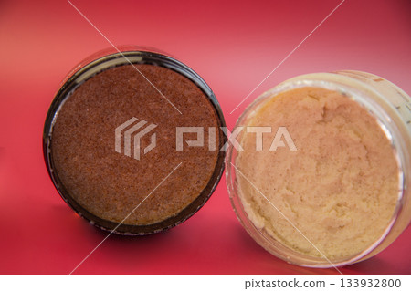 Two jars of body scrub, one brown and one beige, are displayed against a vivid red background, highlighting their unique textures and colors for skincare lovers Two jars of body scrub, one brown and one beige, are displayed against a vivid red background, highlighting their unique textures and colors for skincare lovers 133932800