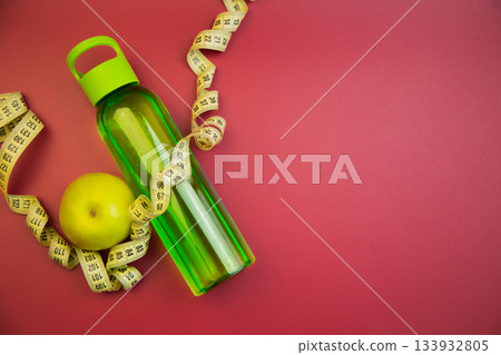 Bright green water bottle alongside a yellow measuring tape and a fresh apple on a vivid red background, representing health and fitness themes for wellness motivation 133932805