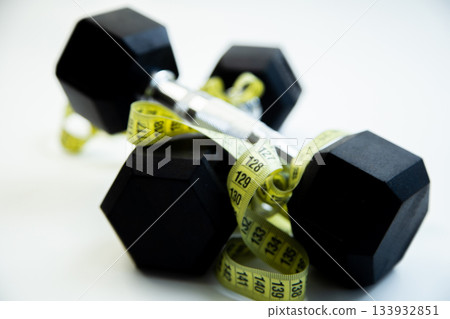 Black dumbbells are intertwined with a yellow measuring tape, representing fitness aspirations and healthy living in a clean, modern workout space Black dumbbells are intertwined with a yellow measuring tape, representing fitness aspirations and healthy living in a clean, modern workout space 133932851