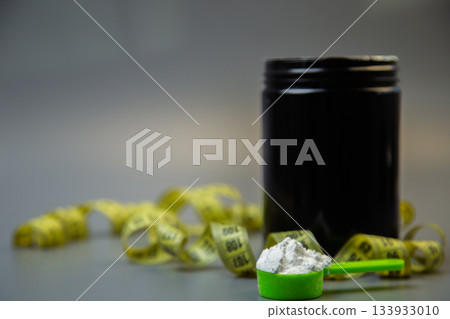 Black jar of protein powder with green scoop filled with white powder, surrounded by yellow measuring tape, emphasizing fitness, nutrition, and health awareness Black jar of protein powder with green scoop filled with white powder, surrounded by yellow measuring tape, emphasizing fitness, nutrition, and health awareness 133933010