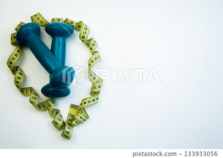 Blue dumbbells encircled by yellow measuring tape form a heart shape on a white background, representing fitness and health in a bright and inspiring atmosphere 133933056