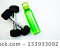 Dumbbells and a green water bottle are arranged on a white surface, symbolizing fitness and hydration, ideal for promoting a healthy lifestyle and exercise routines 133933092