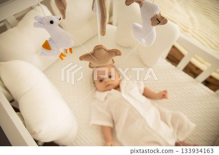 A serene baby resting in a cozy crib beneath soft toys A serene baby resting in a cozy crib beneath soft toys 133934163