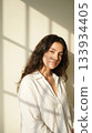 Young woman with flowing dark hair and natural makeup stands in soft sunlight, wearing a white linen shirt and exuding calm elegance against a minimalist wall with gentle shadows. 133934405