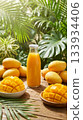 Fresh mango juice in a glass bottle without a label, surrounded by whole and sliced ripe mangoes on wooden table with tropical green leaves and sunlight. 133934406