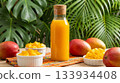 Vibrant arrangement of a glass bottle filled with fresh mango juice, surrounded by ripe mangoes and bowls of diced mango, set on a colorful cloth with tropical green leaves. 133934408