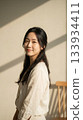 Young Asian woman with long black hair and a gentle smile stands in soft natural light, wearing a light linen shirt in a serene minimalist interior with warm tones. 133934411