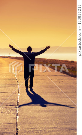 A young man with hands in the air walks along a concrete road and looks at a beautiful orange sunset. Vertical image A young man with hands in the air walks along a concrete road and looks at a beautiful orange sunset. Vertical image 133935213