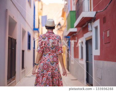 Woman in dress strolls through colorful streets of Spanish coastal town of La Vila Joiosa . sunny winter atmosphere highlights charm of Mediterranean architecture and quiet seaside life, back view 133936288