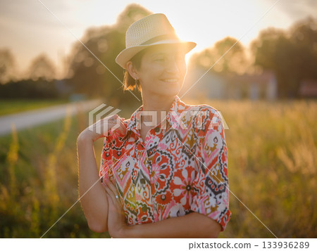 Young woman walking through picturesque European field in late summer. Golden sunlight, lush greenery, and serene rural atmosphere create peaceful countryside scene. 133936289
