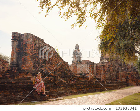 Young female tourist in dress exploring historic park of Ayutthaya, Thailand. Surrounded by ancient temples and statues, she enjoys peaceful atmosphere and rich cultural heritage. 133936341
