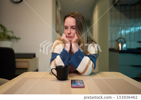 Female resting head on hands at kitchen table with mug and smartphone Female resting head on hands at kitchen table with mug and smartphone 133936462