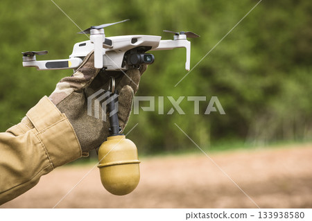 Operator in military colored clothes holds in hand preparing a drone for flight attached grenade Operator in military colored clothes holds in hand preparing a drone for flight attached grenade 133938580