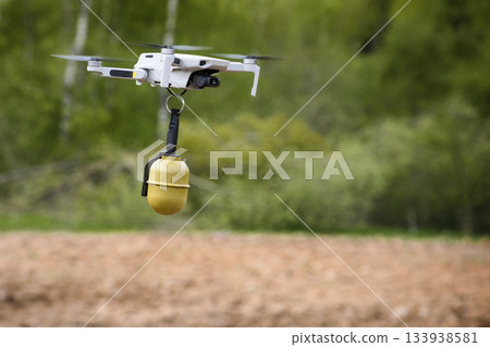 Small Flying unmanned aerial vehicle in flight mode with camera with a grenade attached sideways outdoor green forest plowed field 133938581
