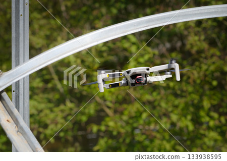 Small Flying Quadcopter Drone in flight mode with camera pointed at viewer outdoor destroyed rebar frame sunny day green background 133938595
