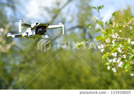 Small Flying unmanned aerial vehicle in flight mode with camera outdoor on sunny day spring time on cherry blossom garden background 133938605