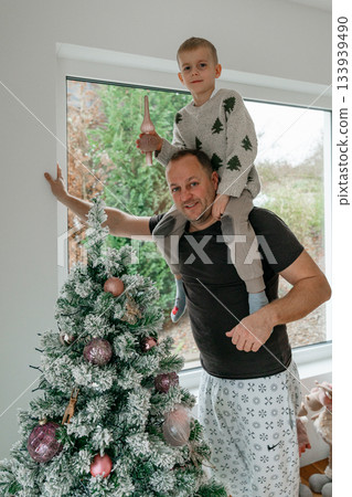 A joyful moment of an adult helping a child decorate a festive Christmas tree. Warm natural light, cozy home atmosphere, winter holiday mood. Candid family scene perfect for seasonal content A joyful moment of an adult helping a child decorate a festive Christmas tree. Warm natural light, cozy home atmosphere, winter holiday mood. Candid family scene perfect for seasonal content 133939490