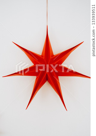 Large red paper star ornament hanging on a clean white wall in soft daylight. Simple and striking Christmas decor perfect for modern interiors, festive styling and seasonal designs Large red paper star ornament hanging on a clean white wall in soft daylight. Simple and striking Christmas decor perfect for modern interiors, festive styling and seasonal designs 133939511