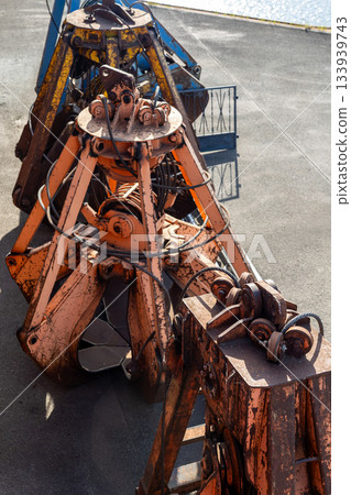 Row of heavy grab buckets rests on concrete surface, showing worn paint, rust textures and complex metal mechanics. Industrial tools create rugged atmosphere with strong sunlight and sharp shadows 133939743