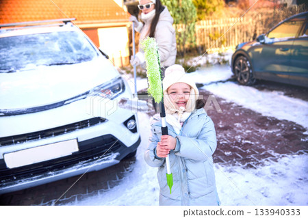 Cute little girl holds snow brush while mom cleans off car in snowy driveway during winter Cute little girl holds snow brush while mom cleans off car in snowy driveway during winter 133940333