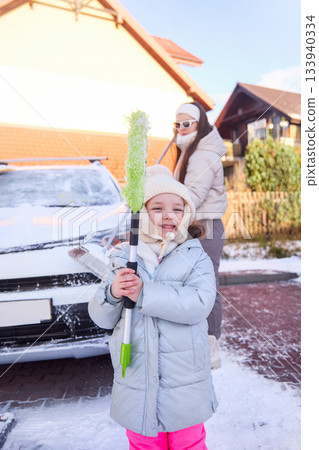 Child holds snow brush while woman clears car on a cold winter day in poland Child holds snow brush while woman clears car on a cold winter day in poland 133940334