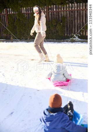 Family fun in Winter sun a day of sledding with mother pulling kids on sleds down snowy hill in winter park 133940344