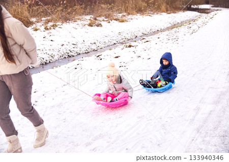 Woman pulling children on plastic sleds through snow-covered field during a Winter day, enjoying a fun activity together during a holiday 133940346