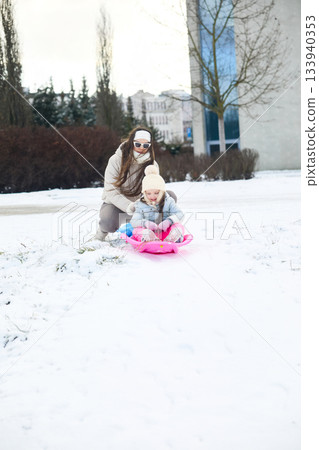 Mother pushes daughter on sled across snow-covered ground near building on winter day 133940353