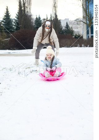 Little girl enjoys a pink sled ride pushed by woman in winter at snowy Park 133940354