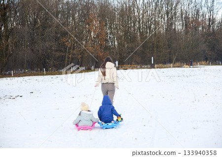 Woman pulls two children on sleds across a snowy field near a bare forest in Winter, enjoying an afternoon activity 133940358
