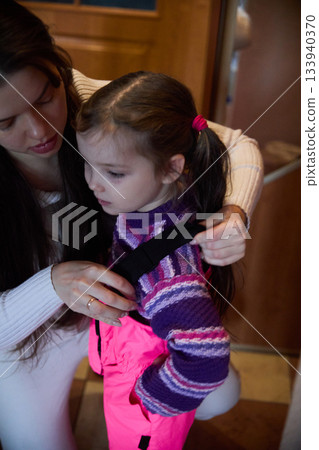Mother helping her young daughter put on a ski suit inside a warm cabin on a winter day, preparing for outdoor activities and family fun Mother helping her young daughter put on a ski suit inside a warm cabin on a winter day, preparing for outdoor activities and family fun 133940370