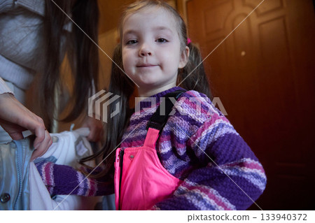 Getting ready for snow day child prepares for outdoor activities with help of guardian near door, dressed in purple sweater and pink snow pants 133940372