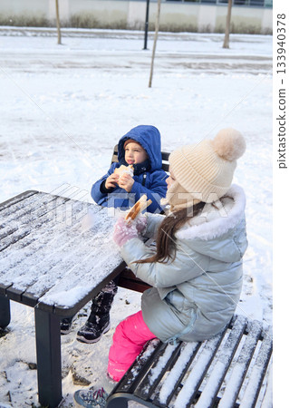 Children eating lunch at a picnic table covered in snow during winter in a park Children eating lunch at a picnic table covered in snow during winter in a park 133940378