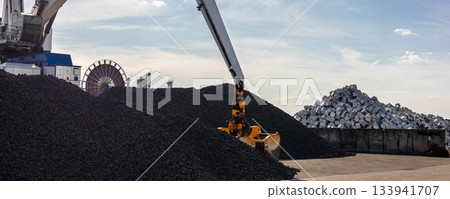 Large industrial crane loads dark coal piles bright sunny sky  backlight background. Metallic blocks and massive equipment storage site. Energy commodity industry global trade logistics business 133941707