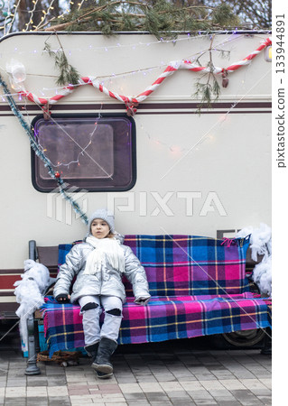 Young child in silver winter coat sits on plaid bench beside decorated camper Young child in silver winter coat sits on plaid bench beside decorated camper 133944891