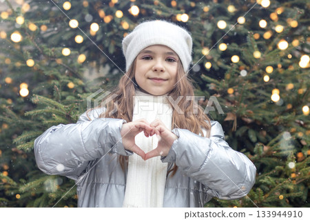 Girl in winter coat and scarf forms a heart with hands in a festive Christmas setting Girl in winter coat and scarf forms a heart with hands in a festive Christmas setting 133944910