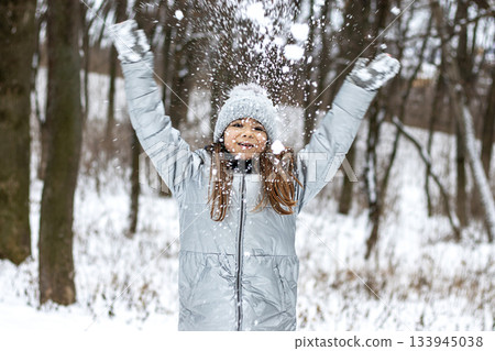 Girl in gray winter coat and knit hat throws snow in a snowy forest 133945038