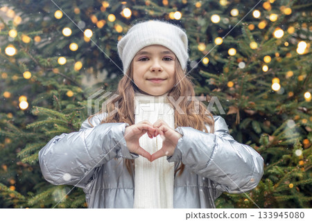Girl in winter coat makes heart sign in front of Christmas tree lights 133945080