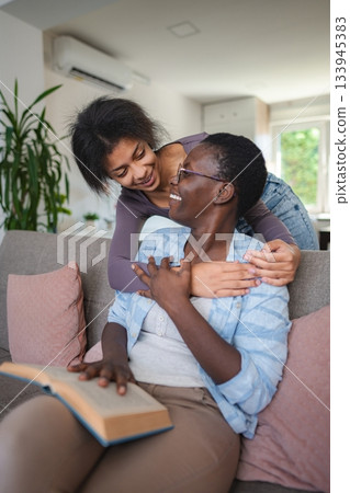 Daughter hugging her mother reading a book on the sofa at home Daughter hugging her mother reading a book on the sofa at home 133945383