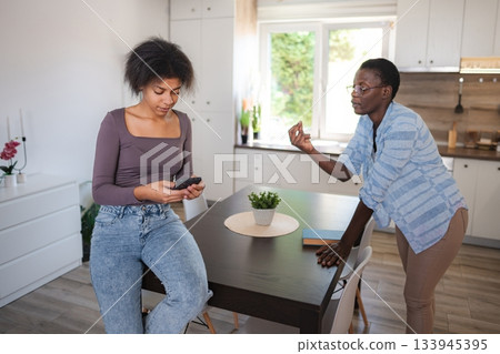 Mother scolding teenage daughter using smartphone in the kitchen 133945395