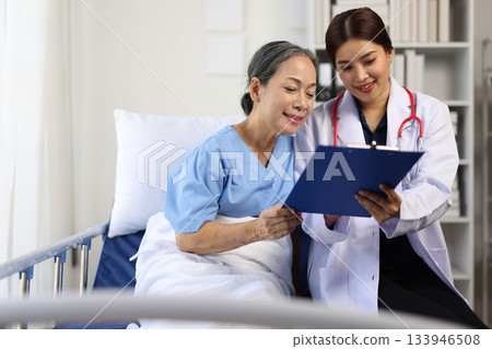 Young female doctor presenting medical results on a clipboard to an elderly female patient, recovering comfortably in a hospital bed. 133946508